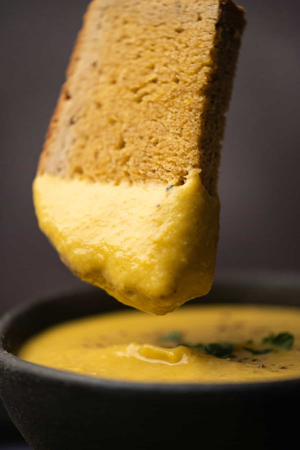 A slice of bread dipping into a bowl of red lentil soup. 