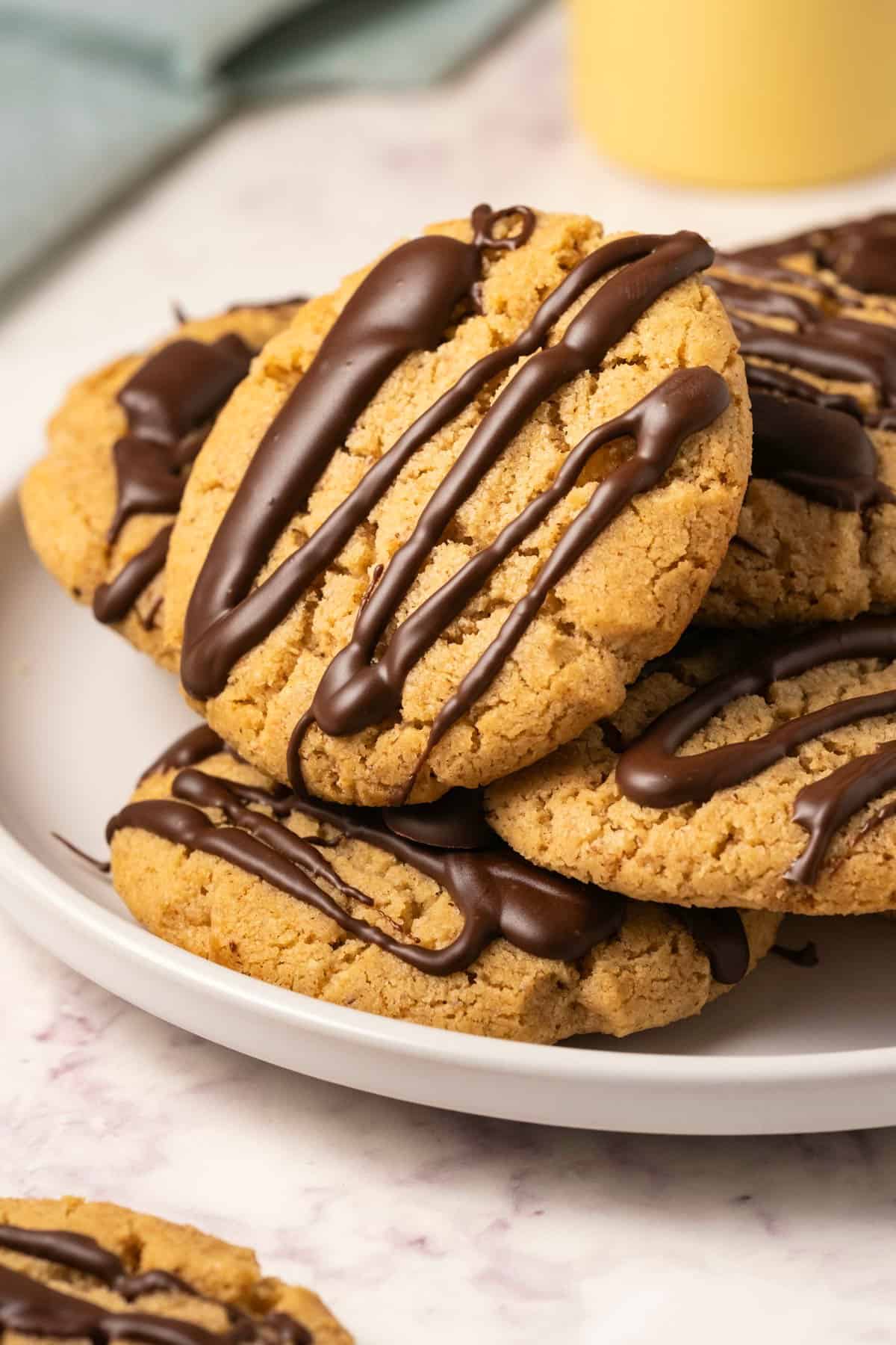 Almond butter cookies topped with a chocolate drizzle stacked on a white plate. 