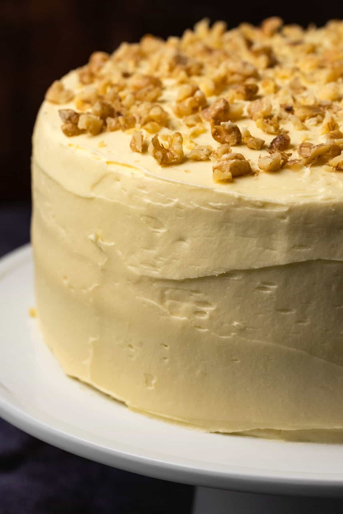 Carrot cake with cream cheese frosting and chopped walnuts on a white cake stand. 