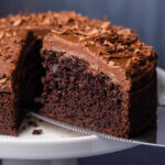 Photo of a cake on a white cake stand with one slice cut and ready to serve.