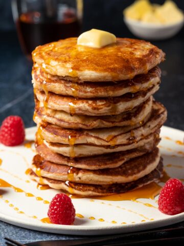 Stack of pancakes topped with butter and drizzled with syrup, on a white plate.