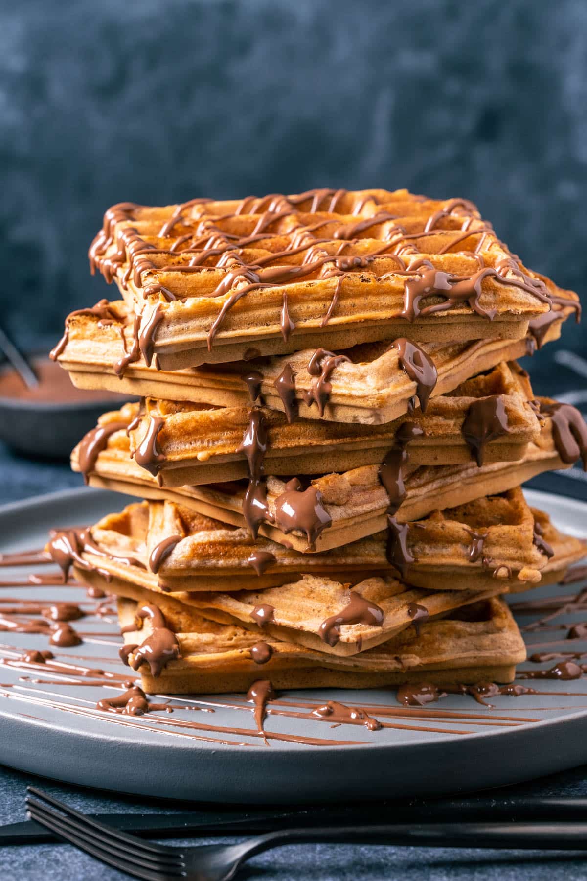 Stack of nutella waffles on a gray plate, drizzled with melted nutella. 