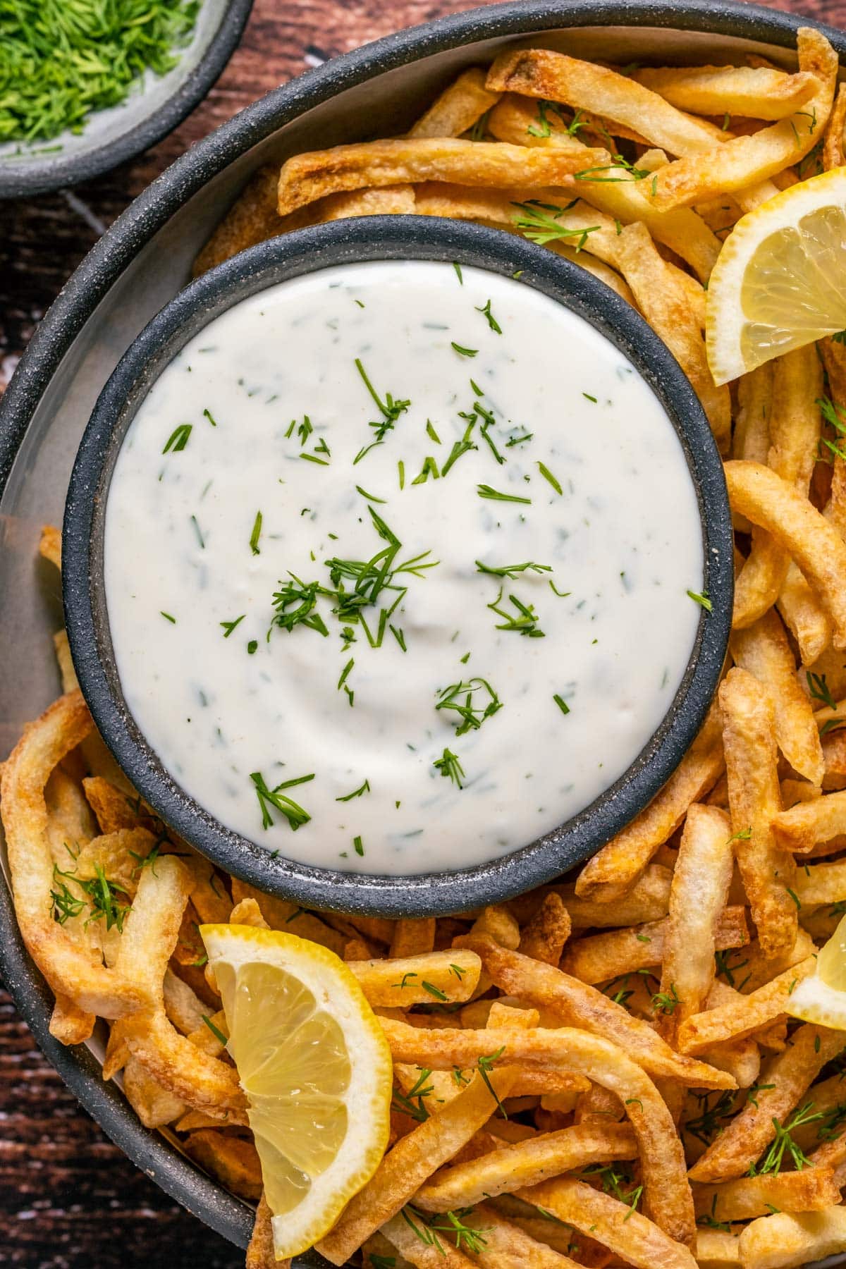 Lemon dill aioli topped with a sprinkle of dill on a plate with chips and lemon slices. 